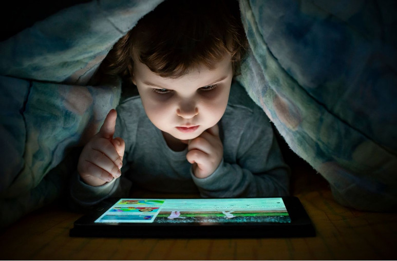 Enfant sous la couette entrain de jouer à la tablette. Un enfant sur un écran